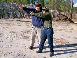 NRA gun safety rule 1: Always keep the gun pointed in a safe direction. Joe Katz is showing Janet Katz the safe direction on this shooting range. Note the directly rooftop behind them, the low berm to the side, and the driveway behind it. Those directions are not safe.